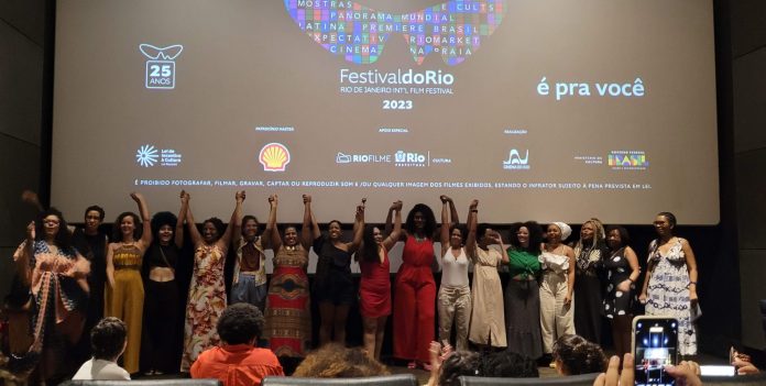 Elenco e equipe do filme Corpos Invisíveis dentro do cinema da Estação Net Gávea levantam os braços uma das outras em sinal de vitória. Estão abaixo da grande tela de cinema que tem escrito Festival do Rio.